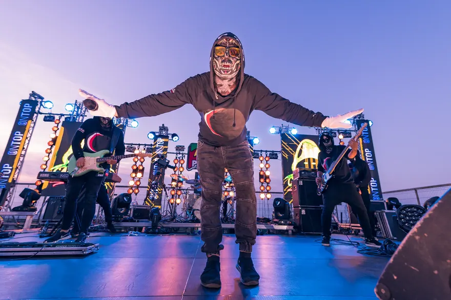 Solist mascat cu brațele deschise aproape de marginea scenei unui festival exterior