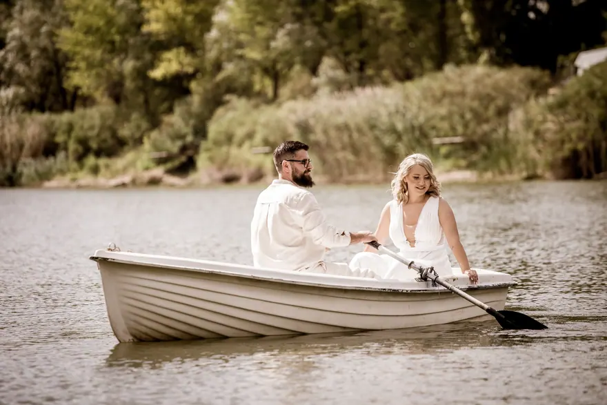Cuplu îmbrăcat în alb într-o barcă mică pe lac, cu malul împădurit în fundal