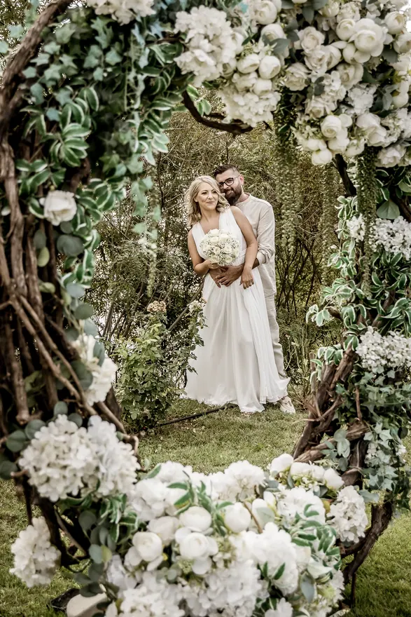 Cuplu în ținute de nuntă fotografiat printre flori albe și frunziș verde